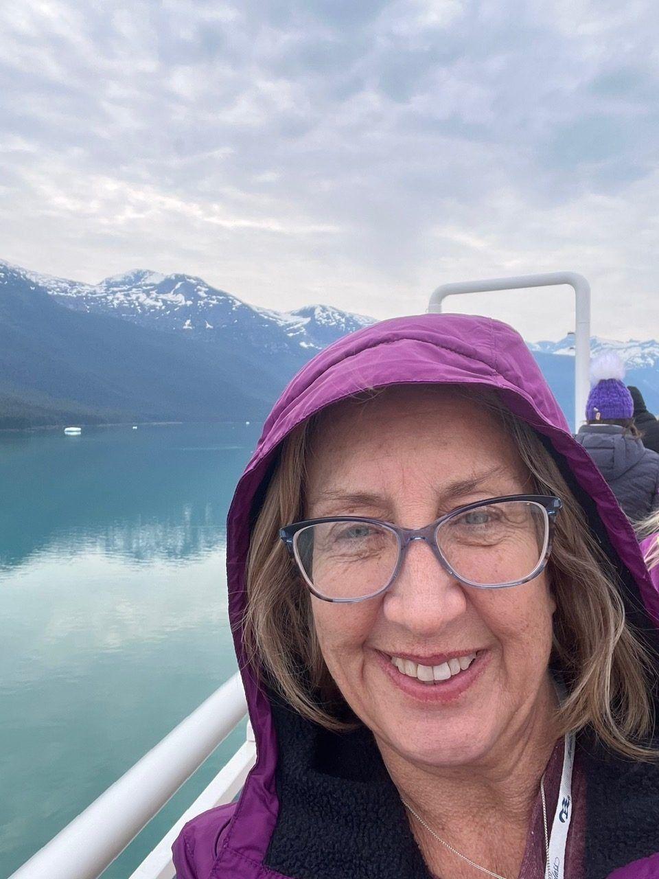 Woman in a purple hooded jacket smiles on a boat with snowy mountains and water behind her.
