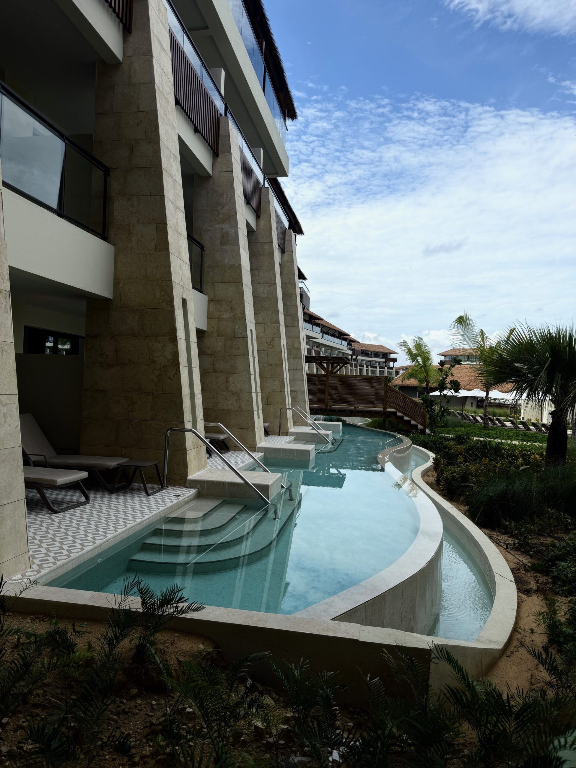 Curved resort pool beside a modern building, with blue water and lounge area overlooking greenery.
