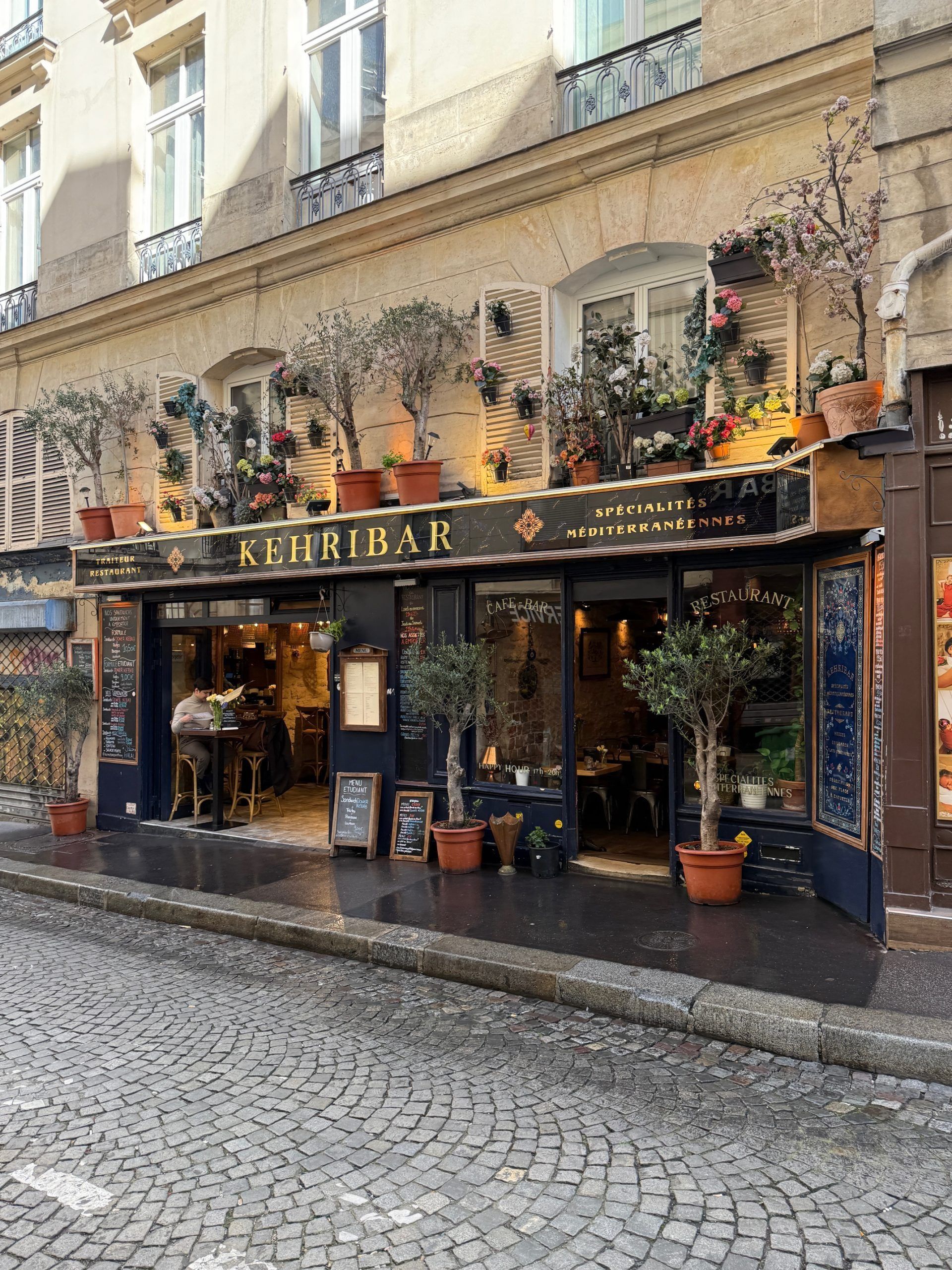 Flower-covered café storefront on a cobblestone street with potted plants and large windows