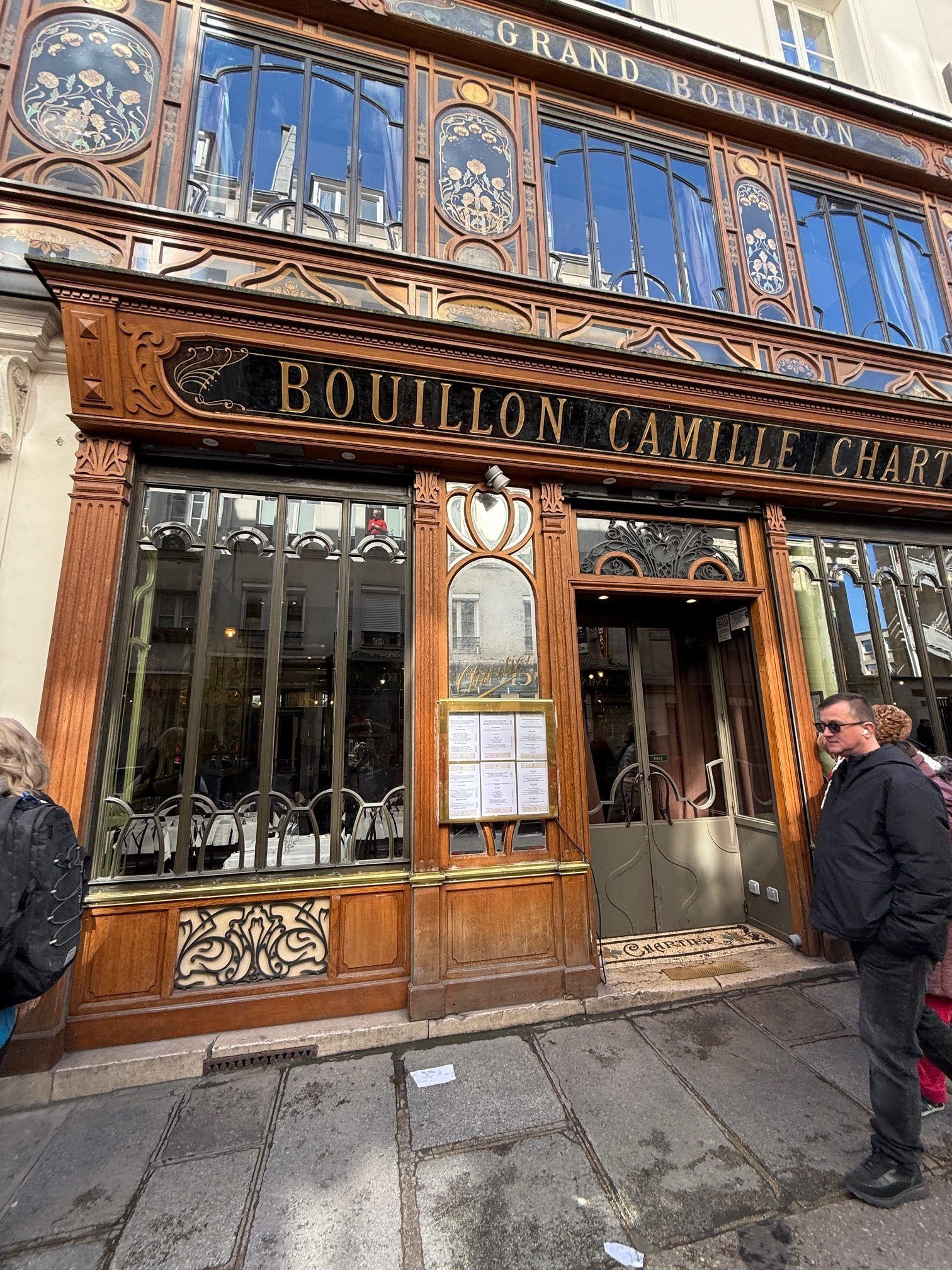 Ornate storefront with gold lettering and blue mosaic windows on a wet street; two people stand outside.