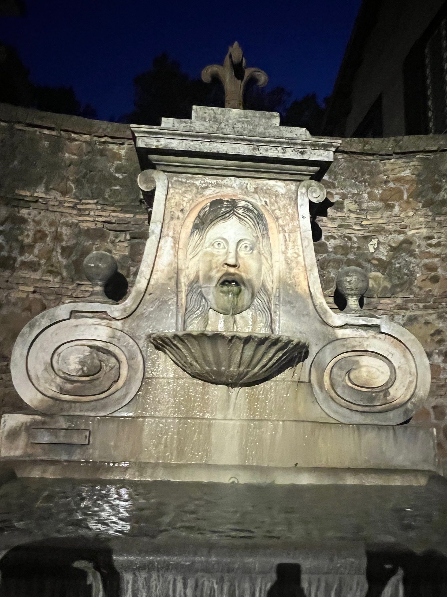 Stone fountain with a carved face and shell basin against a stone wall at night