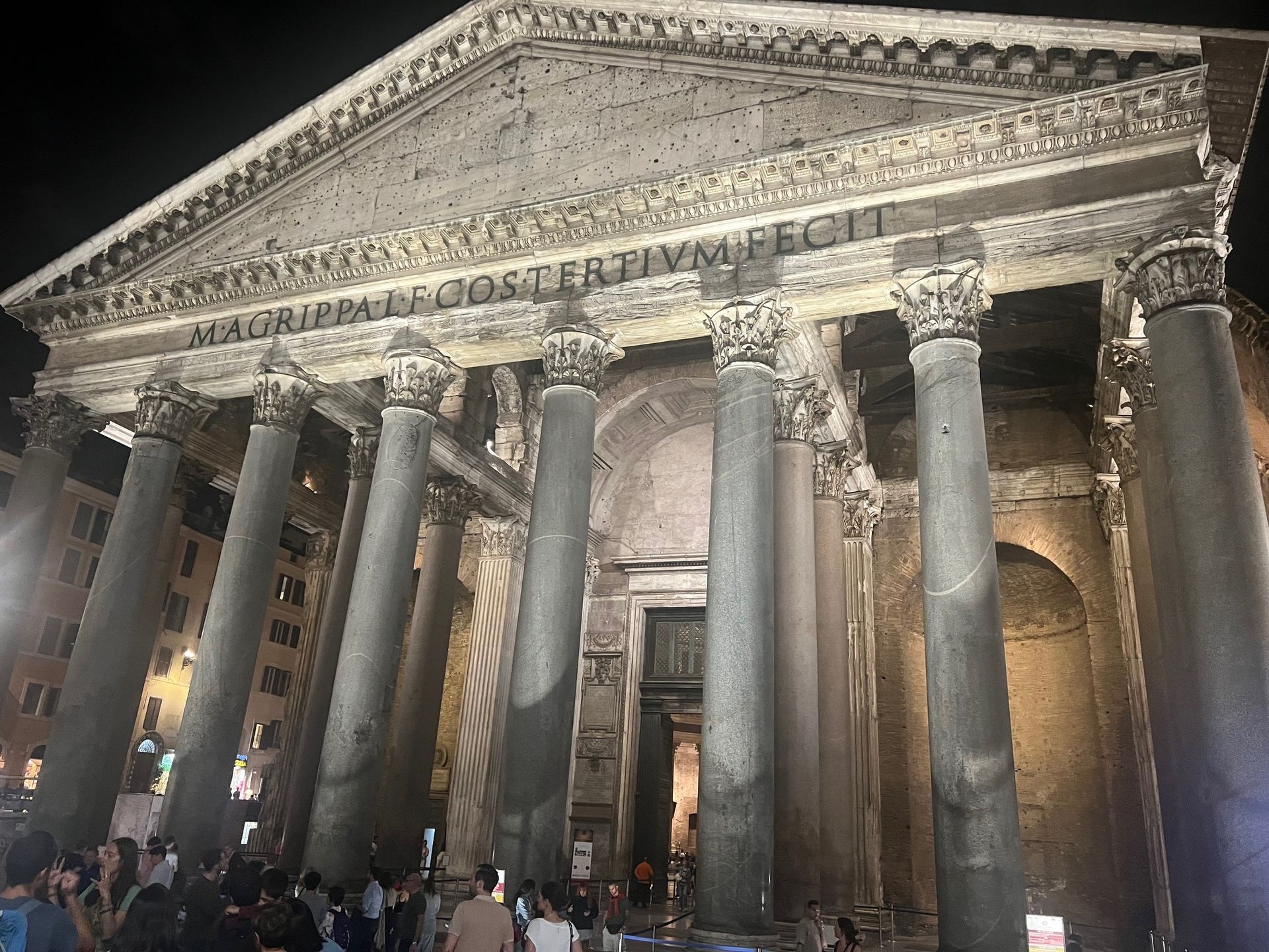 Illuminated ancient Roman temple with towering columns at night, likely the Pantheon, with visitors below