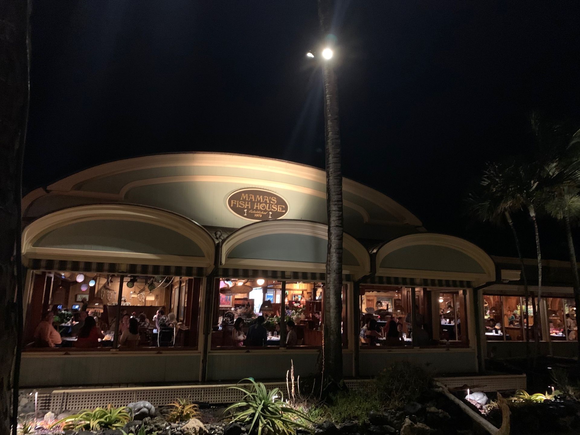 Nighttime view of a lit restaurant with large arched windows and people dining inside