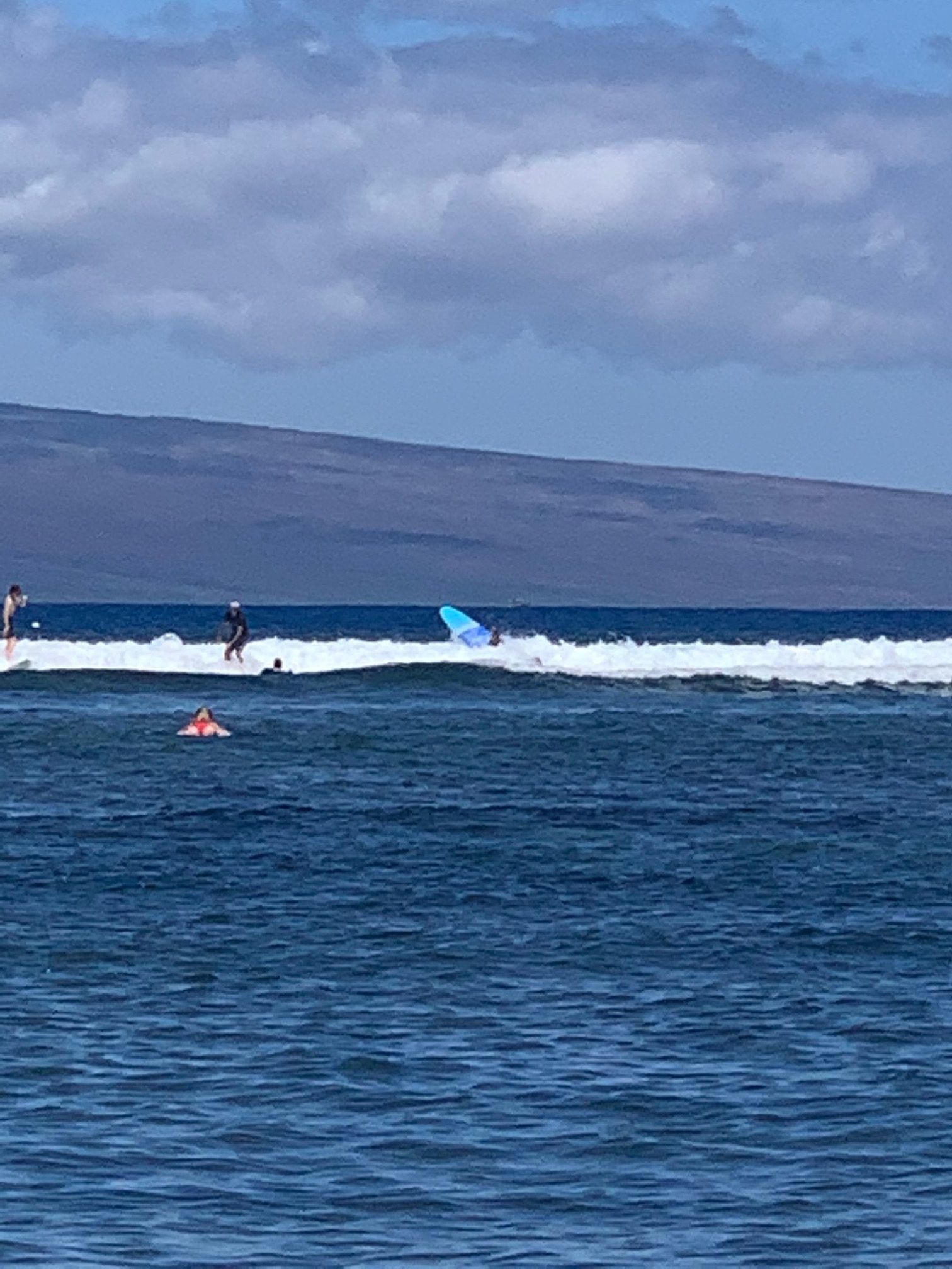 Surfers on blue ocean waves near a distant shoreline under a cloudy sky