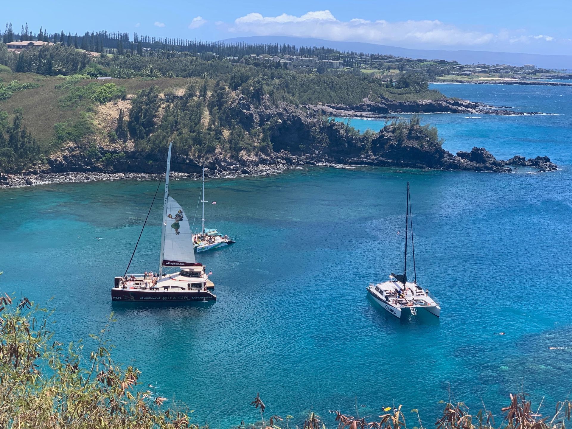 Two sailboats on bright blue water near a rocky coastline under a clear sky
