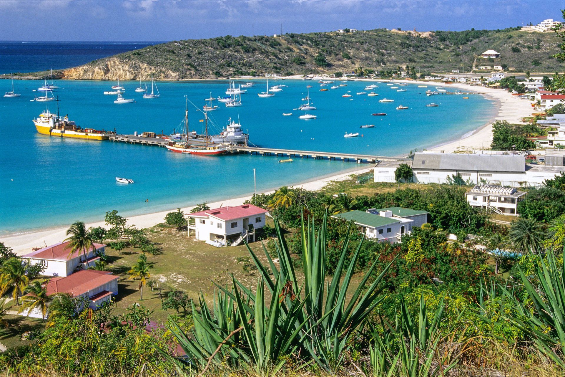 Turquoise bay with boats, a sandy beach, and hillside buildings under a clear blue sky
