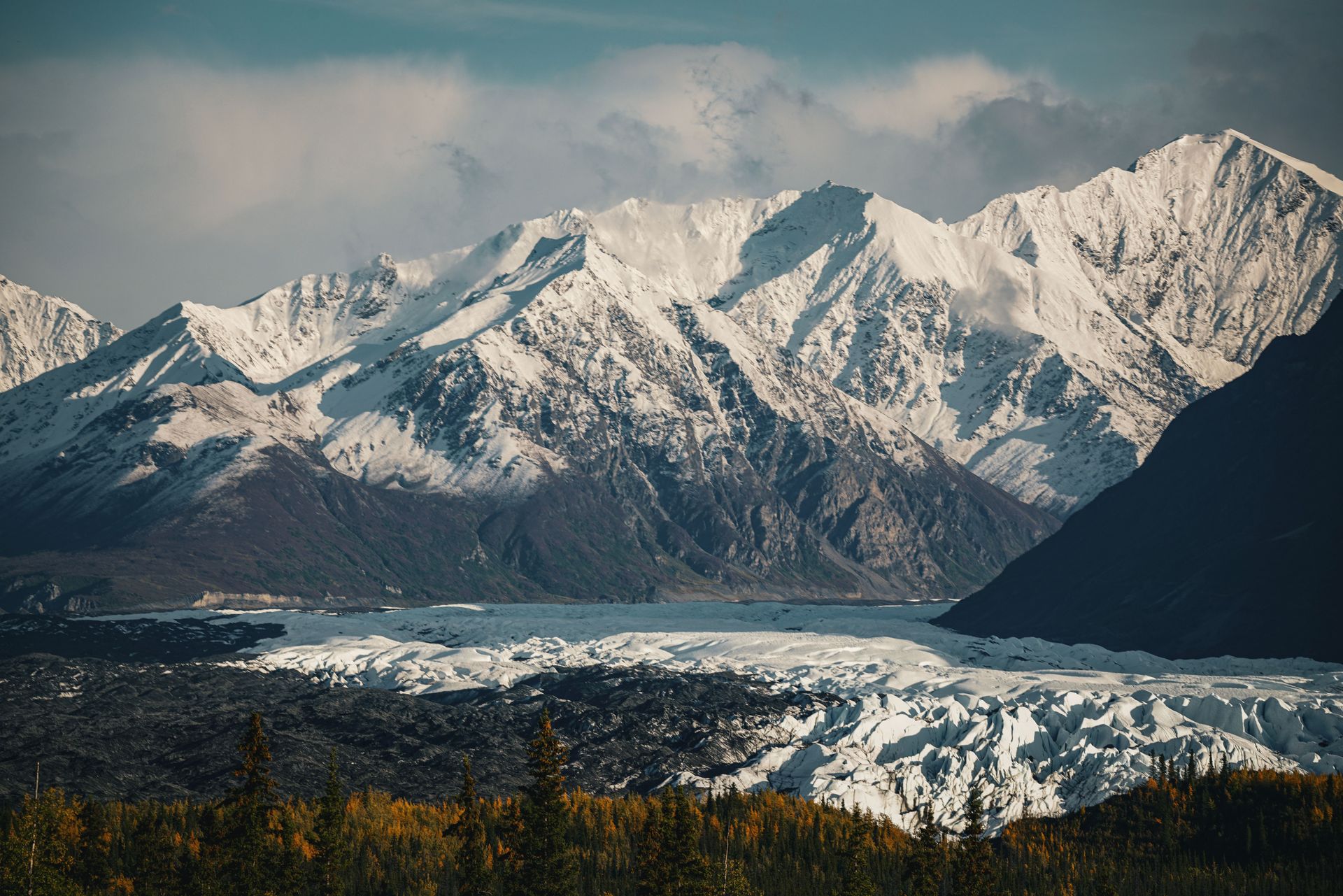 Snowy mountain range above a forested valley under a cloudy blue sky