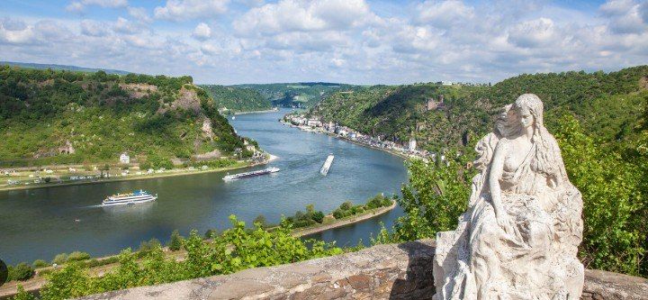 Statue overlooking a wide river valley with boats, green cliffs, and a bright cloudy sky