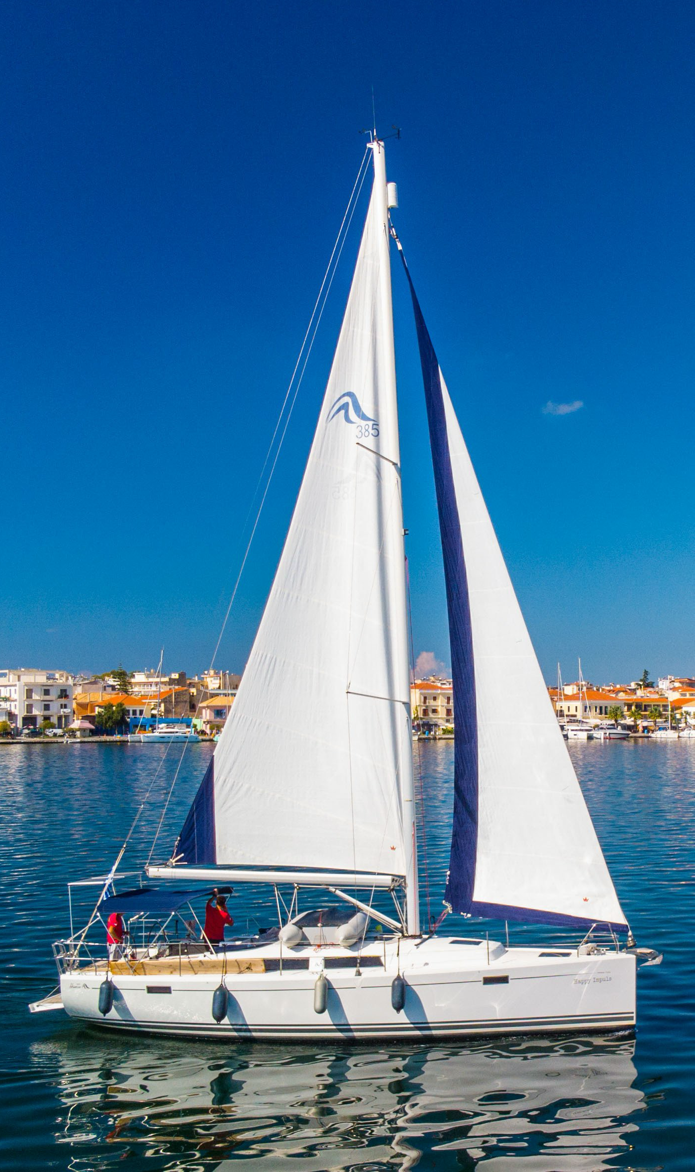 White sailboat with a blue sail cruising on calm water at dusk, city lights in the background