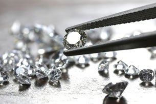 Tweezers holding a clear gemstone above scattered diamonds on a reflective surface