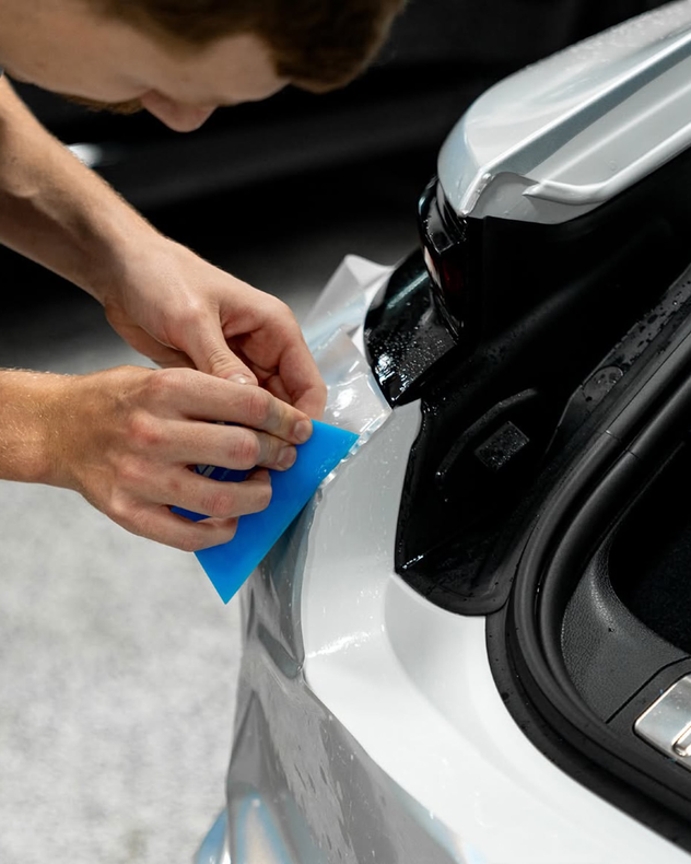 Hands using a blue squeegee to apply clear film on a car hood.