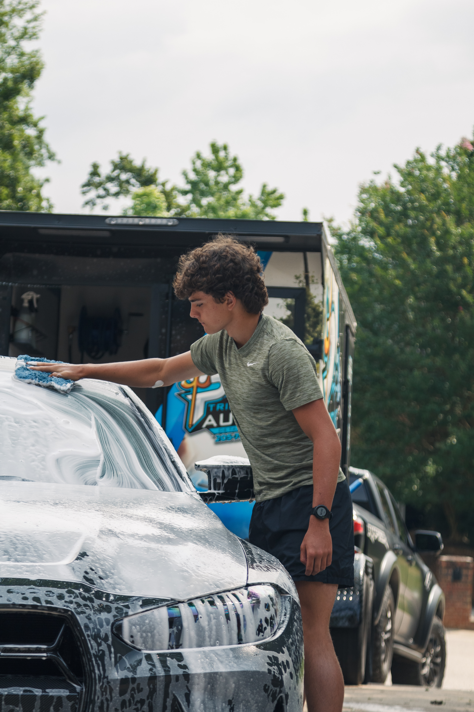 Person washing a white car outdoors with soap suds, holding a sponge beside a truck.