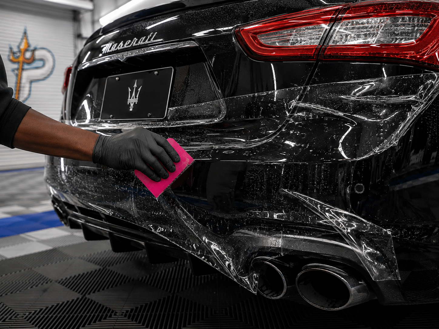 Gloved hand cleaning a black sports car’s rear bumper with a pink cloth in a car wash/detailing bay