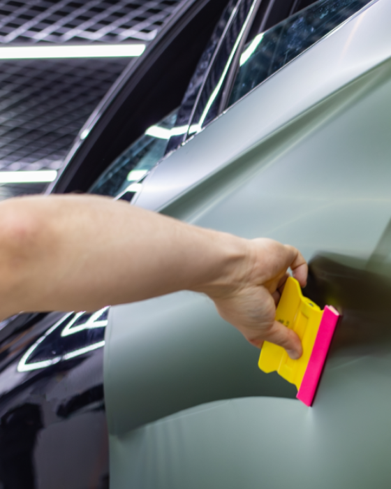 Hand holding a pink-and-yellow squeegee on a car window, cleaning the surface