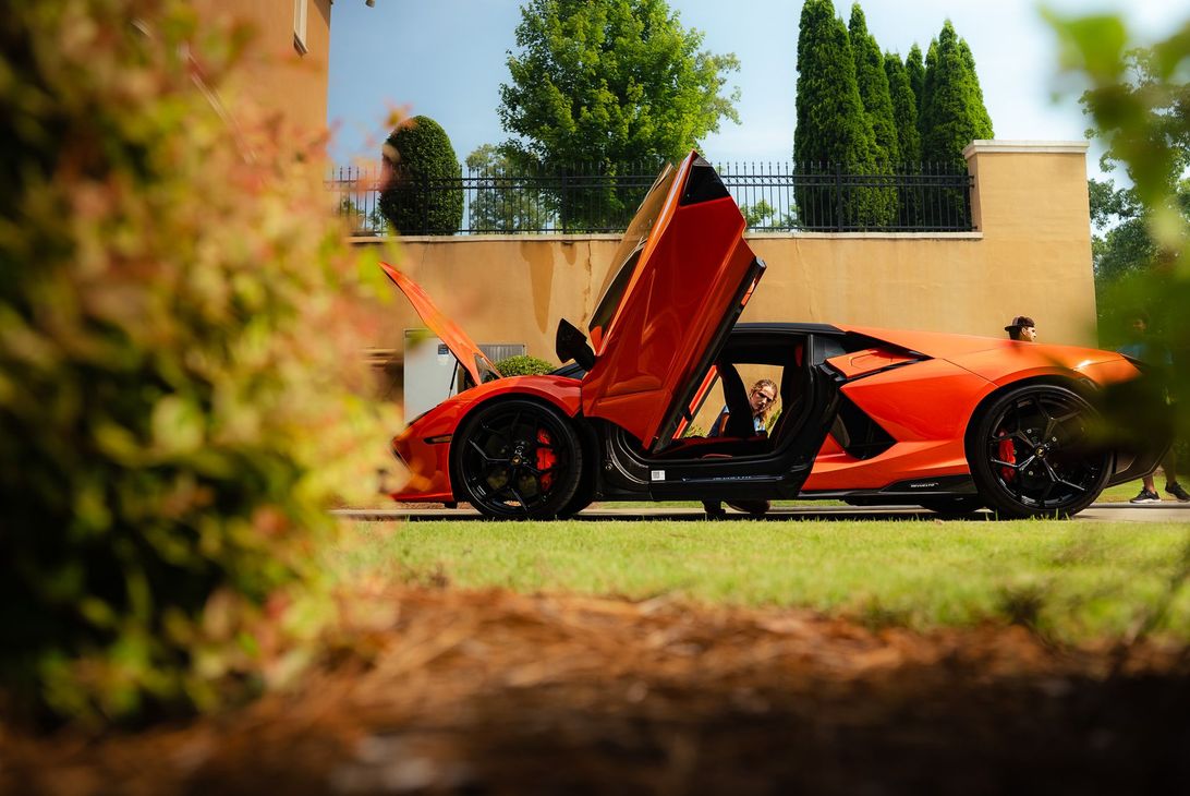 Orange sports car with gullwing doors open in a grassy driveway, seen through blurred foliage in the foreground