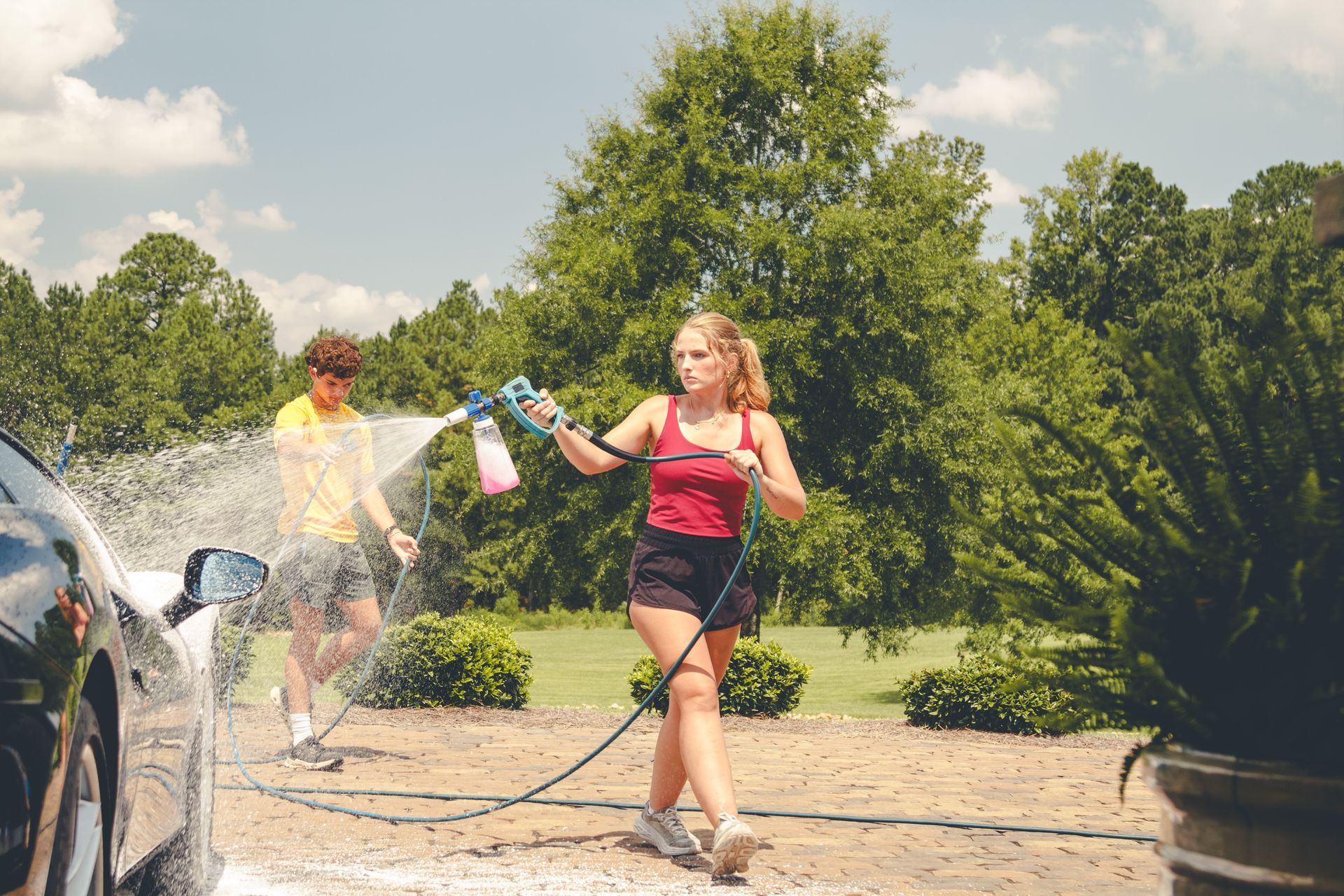 People spraying water outdoors on a sunny day, with a woman in a red top walking through the spray