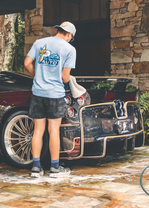 Person washing a black sports car in a driveway, standing beside the front bumper with a hose.