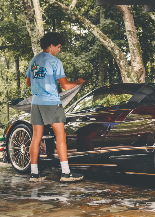 Person standing beside a black sports car in a tree-shaded driveway, holding a phone.