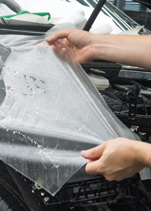 Hands peeling a clear protective film from a gray car hood.