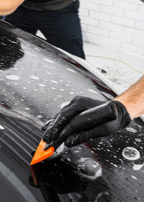 Gloved hand applying a clay bar to a black car hood covered in soap suds