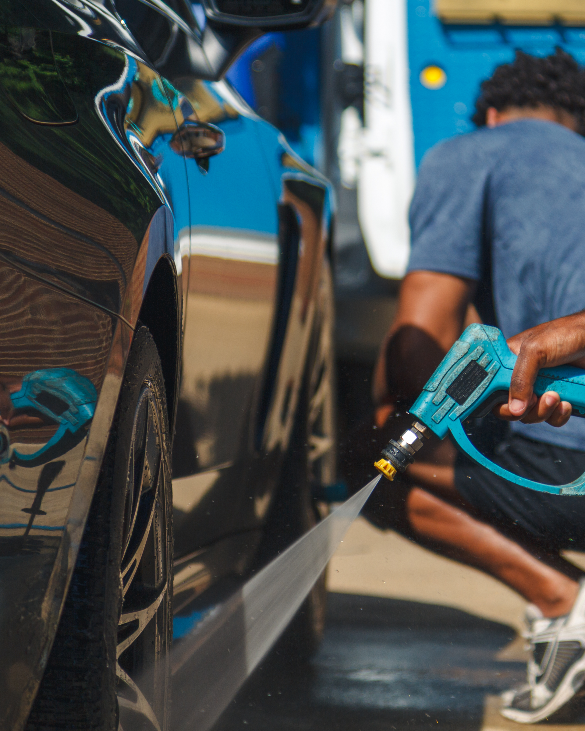 Person washing a blue car with a spray nozzle outdoors