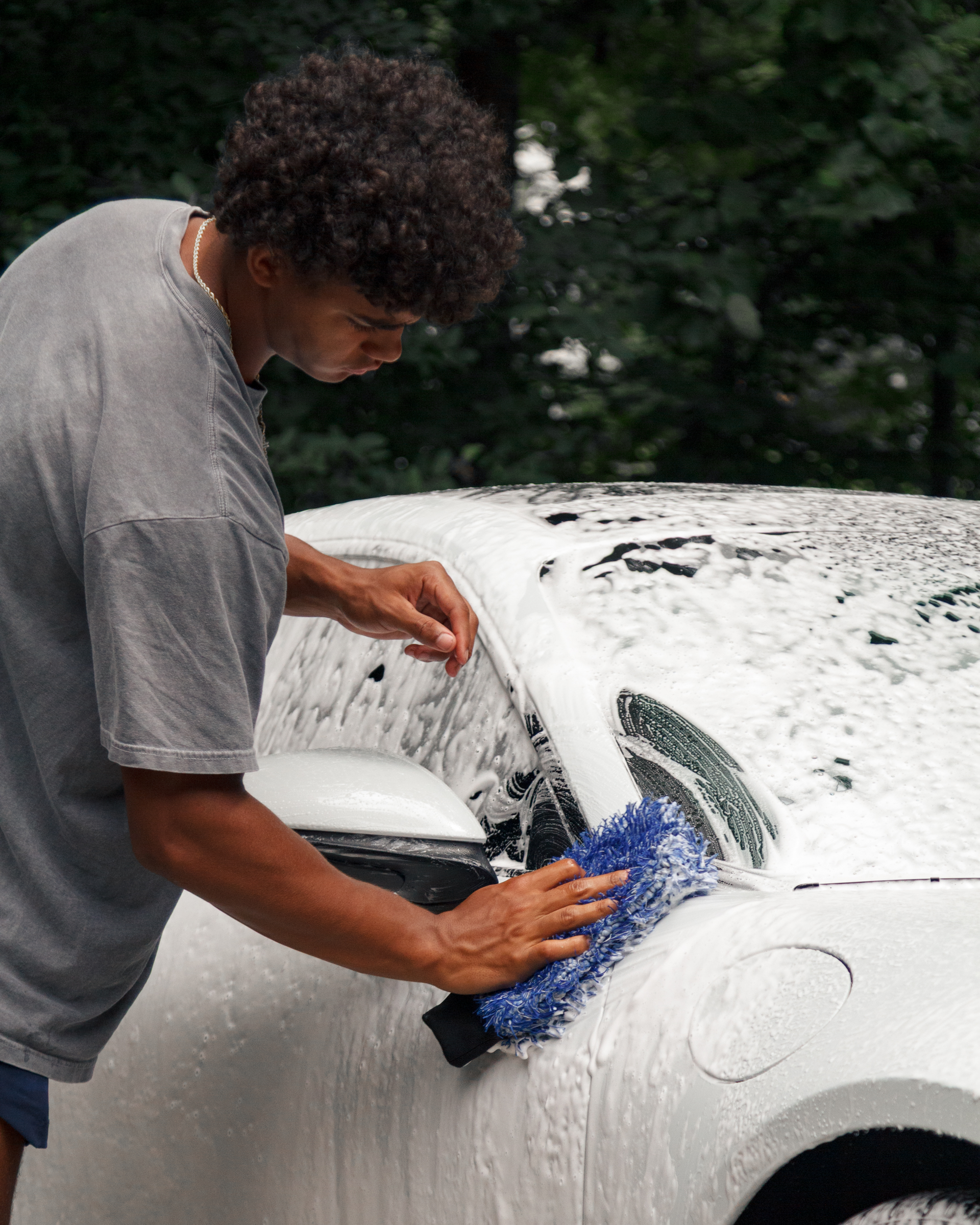 Person washing a white car with a blue sponge, covered in soap suds.