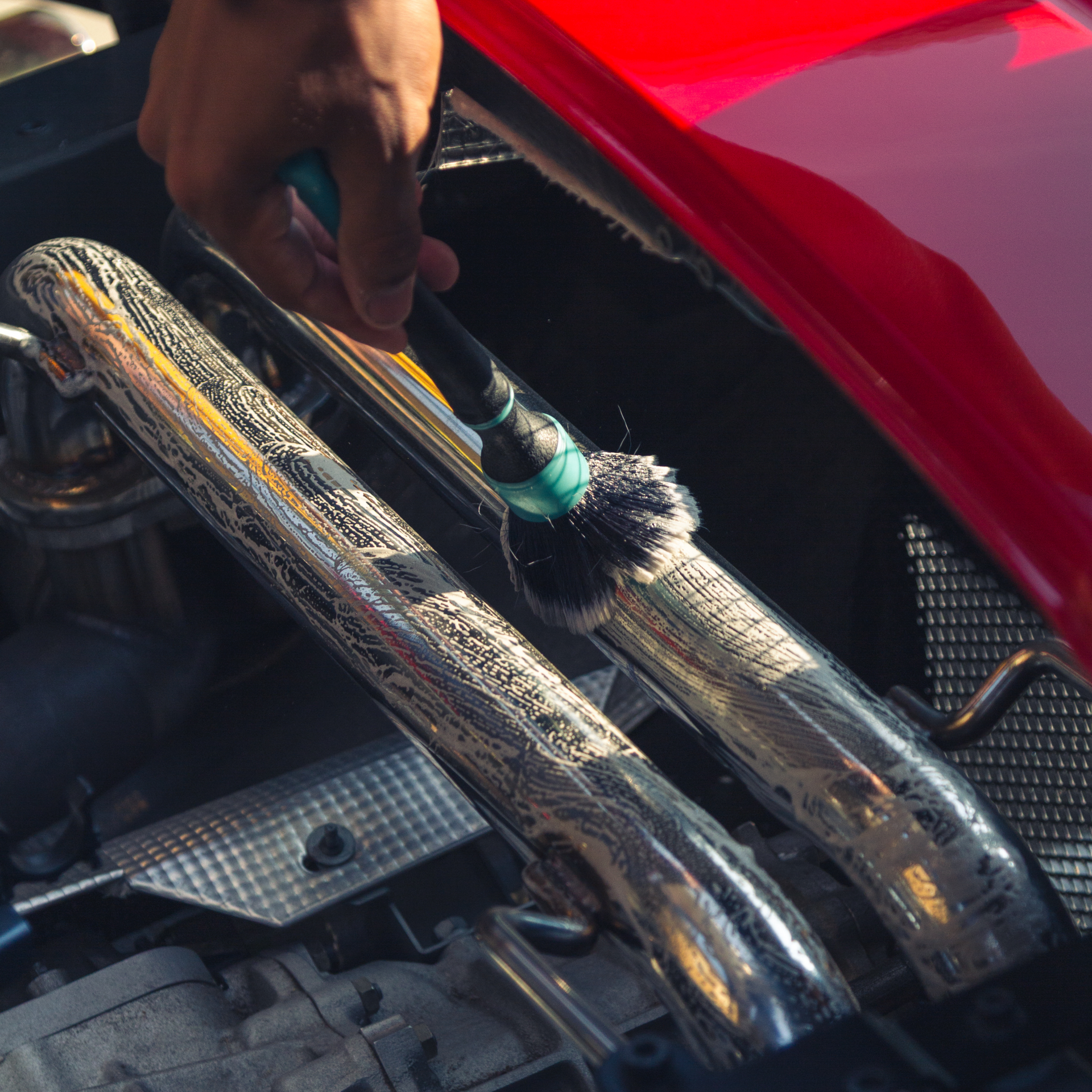 Person holding a toothbrush near a red car engine, cleaning a metal part with a gloved hand.