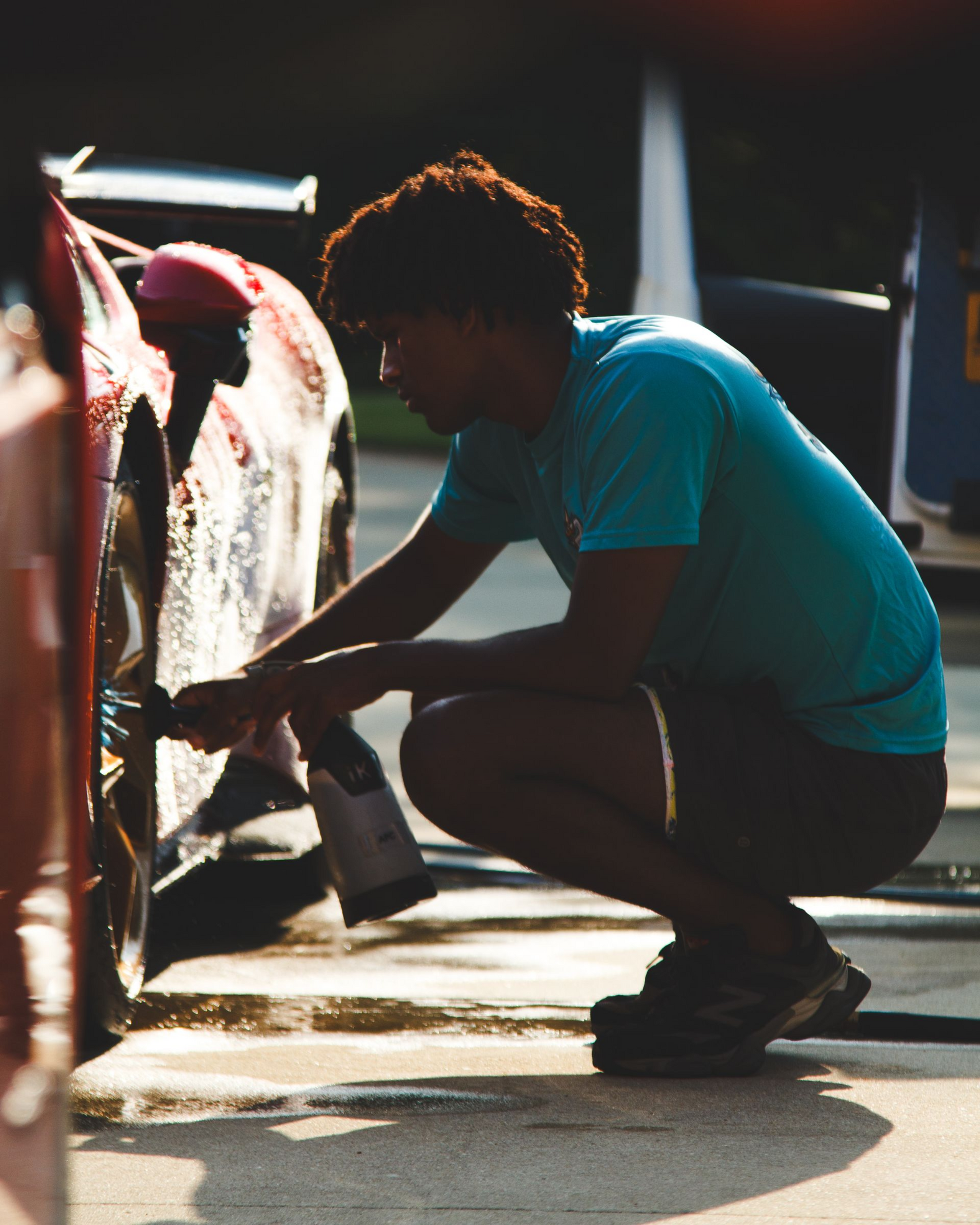 Person crouching and washing a car wheel with a cloth in bright sunlight