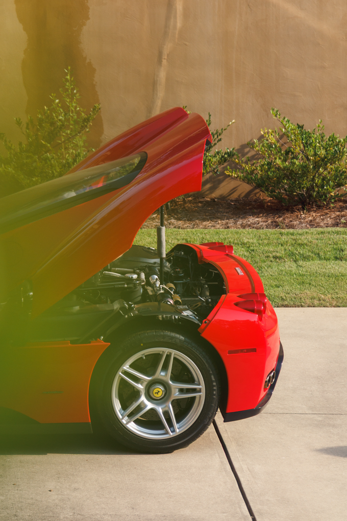 Red sports car with rear engine cover open, parked in a driveway beside a beige wall and greenery.