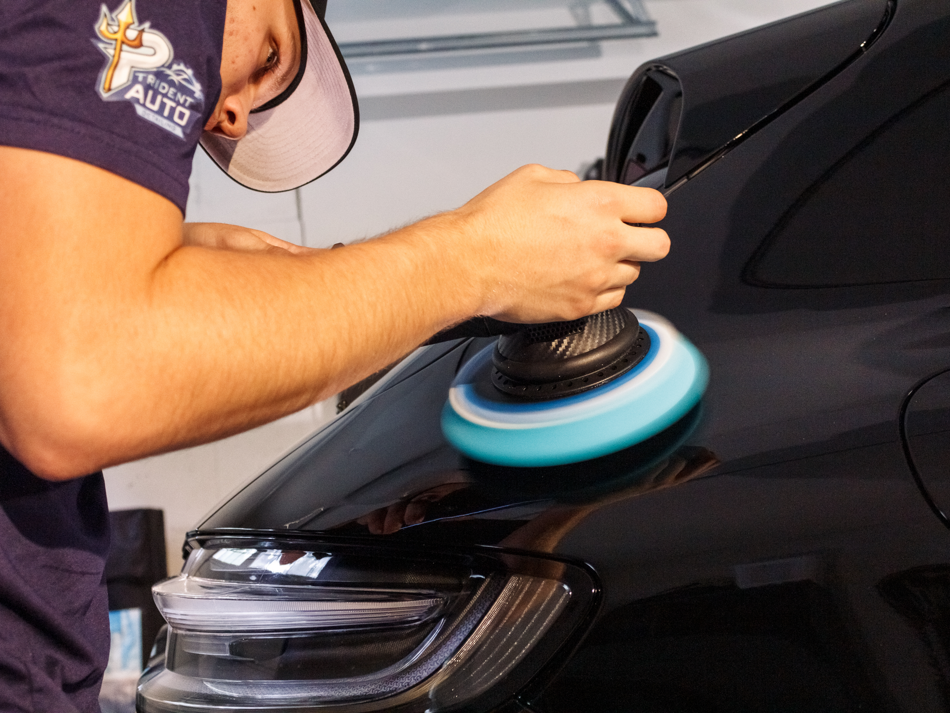 Person polishing a black car’s hood with a blue buffer in a garage