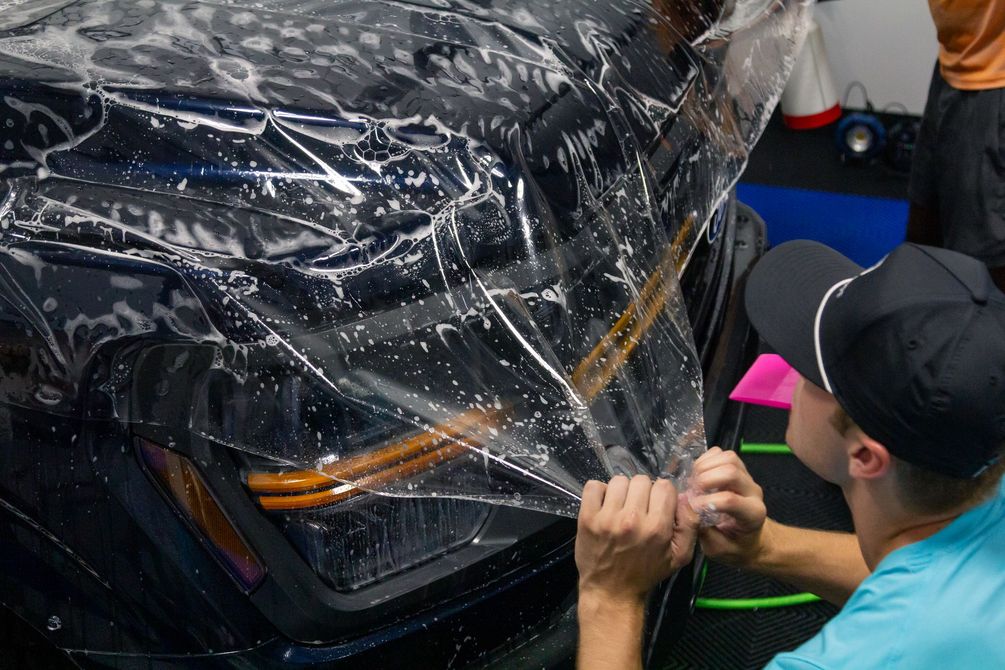 Person applying clear protective film to the front of a black car outdoors.