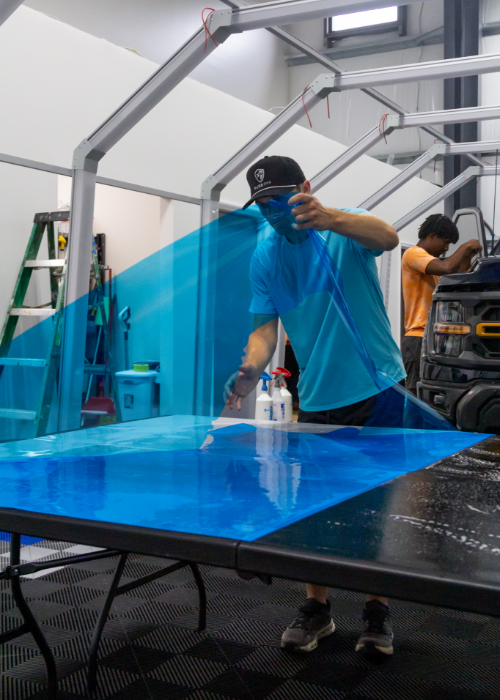 Person in a workshop stretching blue mesh over a table frame.
