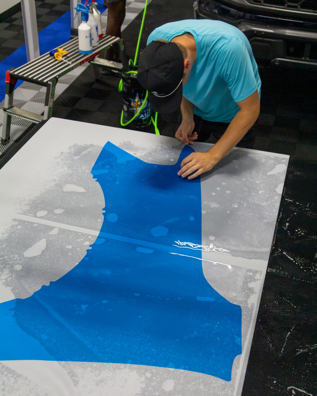 Person crouches over a large gray sheet, applying bright blue material in an industrial workspace.
