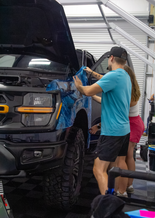 Person applying blue masking tape to a black truck in a garage-like workshop.