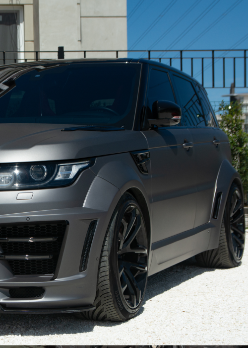 Matte gray SUV with black rims parked outdoors by a building and fence.