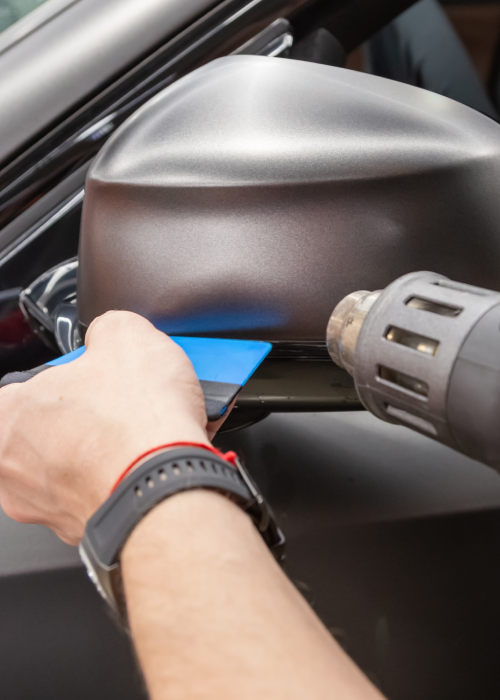 Hands apply blue tape to a car side mirror with a heat gun nearby