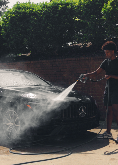 Person pressure-washing a black car in a driveway, with water spraying across the hood.