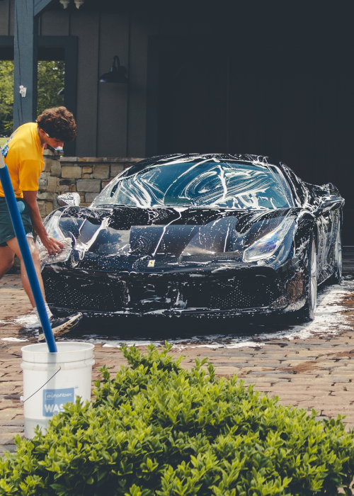 Person washing a black sports car with soap in a driveway outside a house