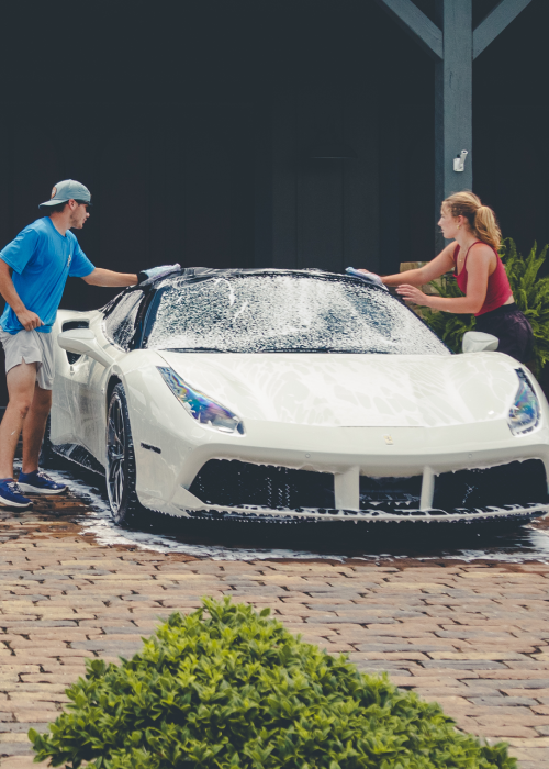 Two people washing a white sports car outdoors with soap and water
