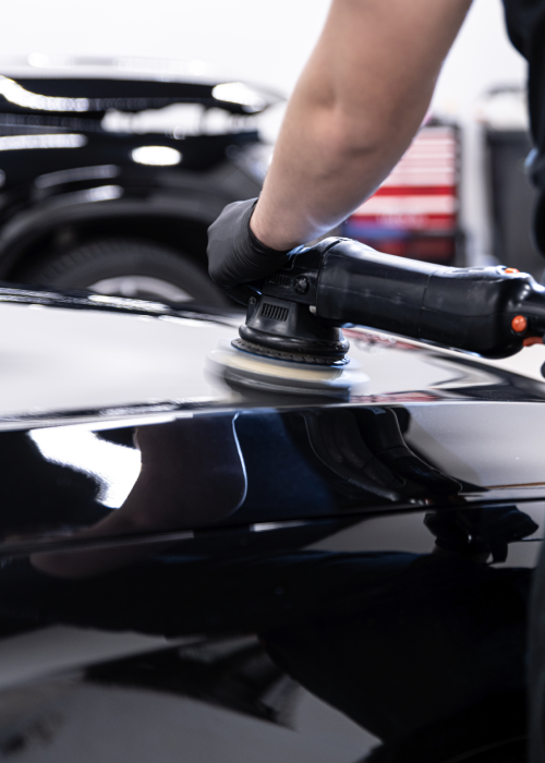 Person polishing a black car hood with a handheld buffer in a garage