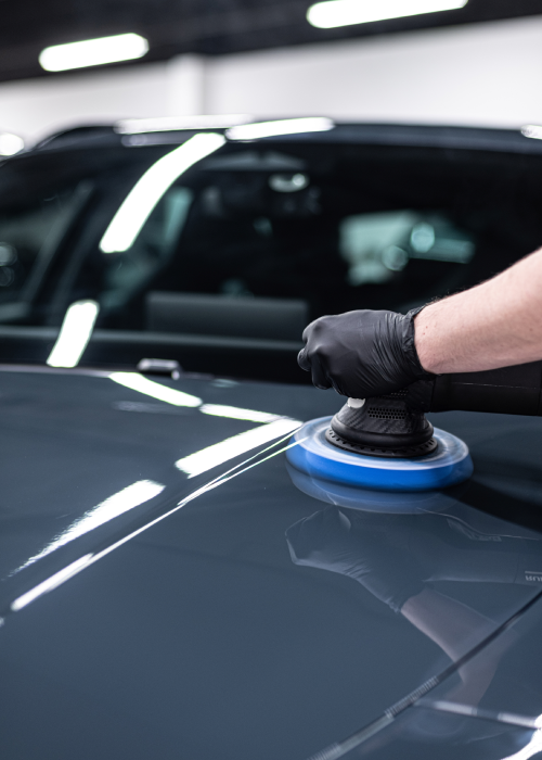 Gloved hand polishing a black car hood with a blue buffer pad in a garage