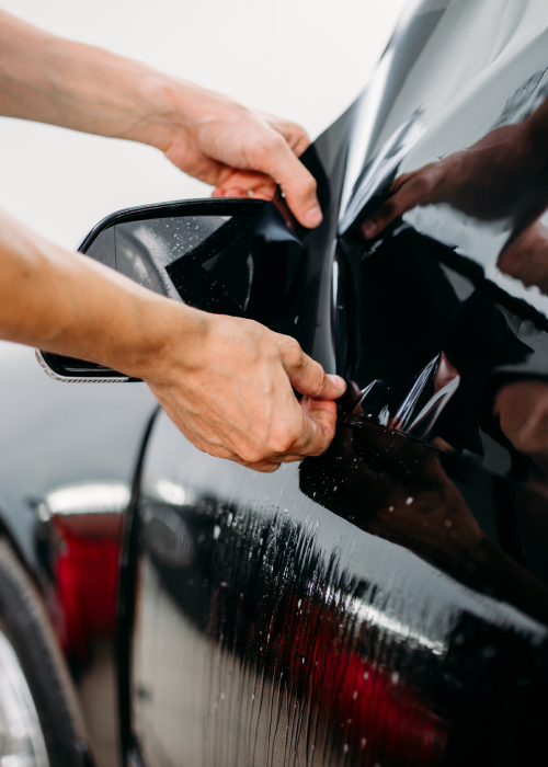 Hands applying vinyl wrap to a black car door in a workshop
