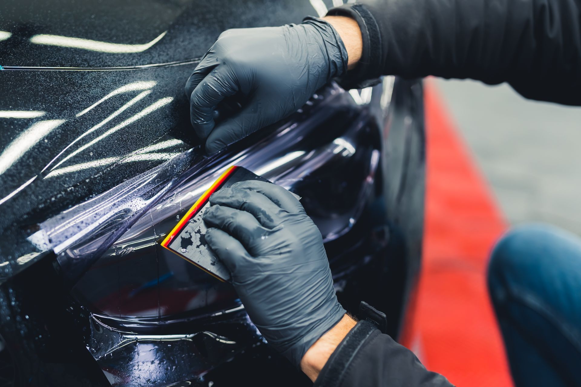 Gloved hands applying orange tape to a black car’s headlight in a workshop