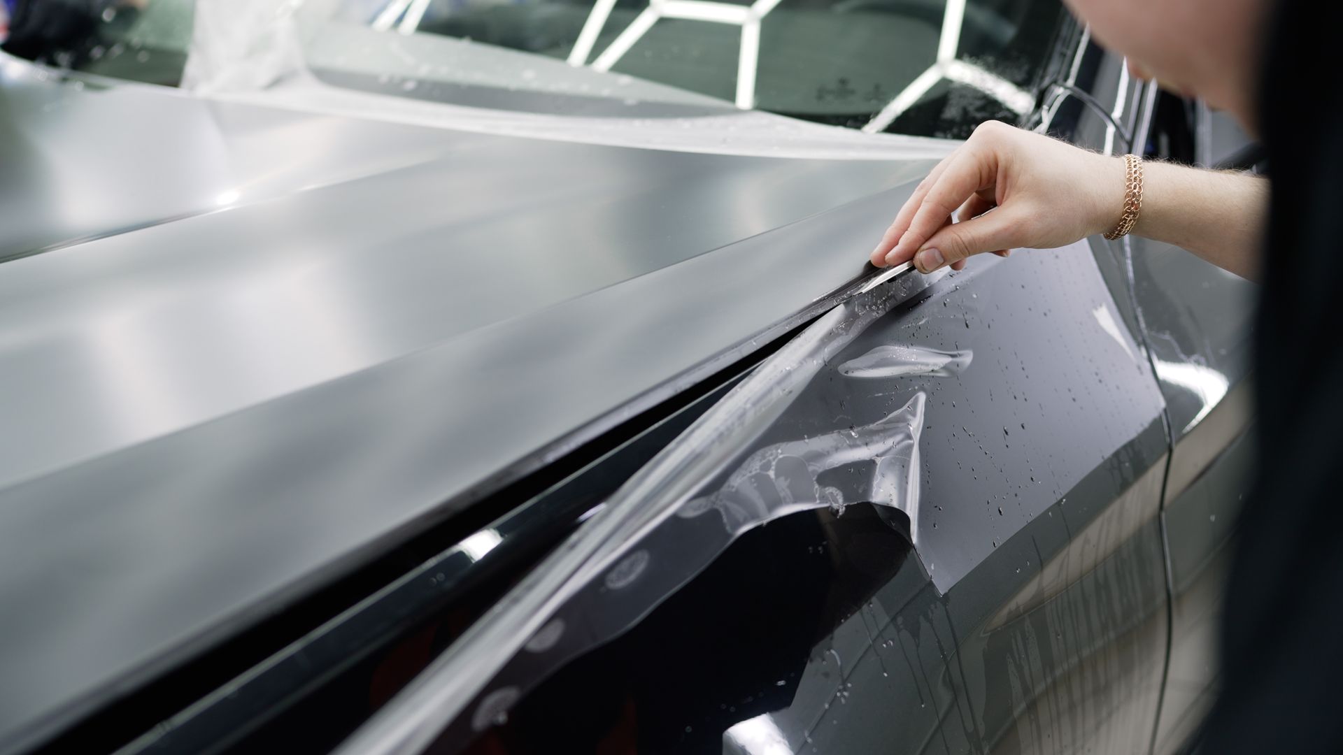 Hand peeling protective film from a glossy black car hood in a workshop
