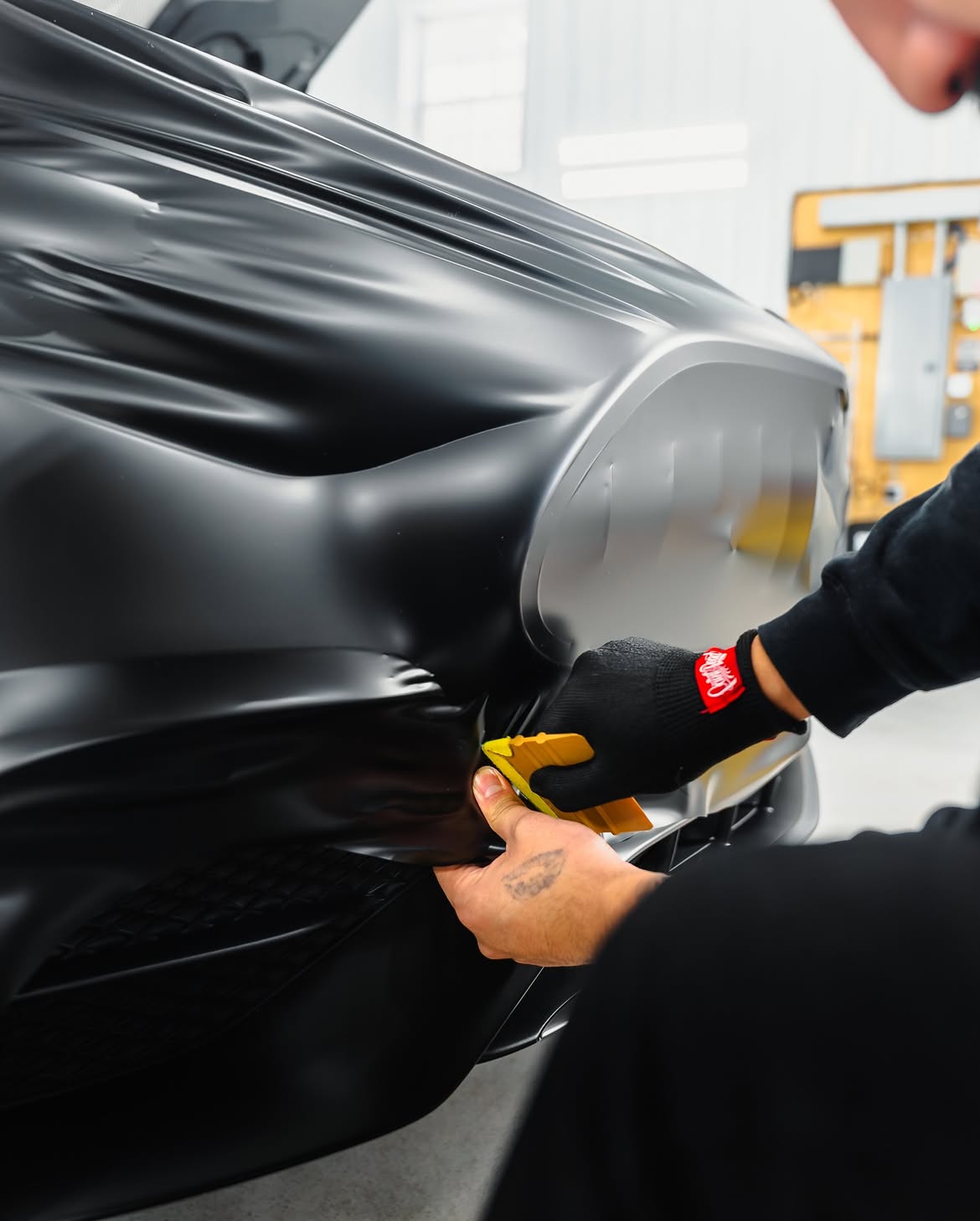 Person applies yellow tape to a black car wrapped in glossy film in a workshop