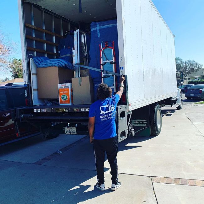 Worker in blue shirt loading boxes into a white moving truck on a sunny street.