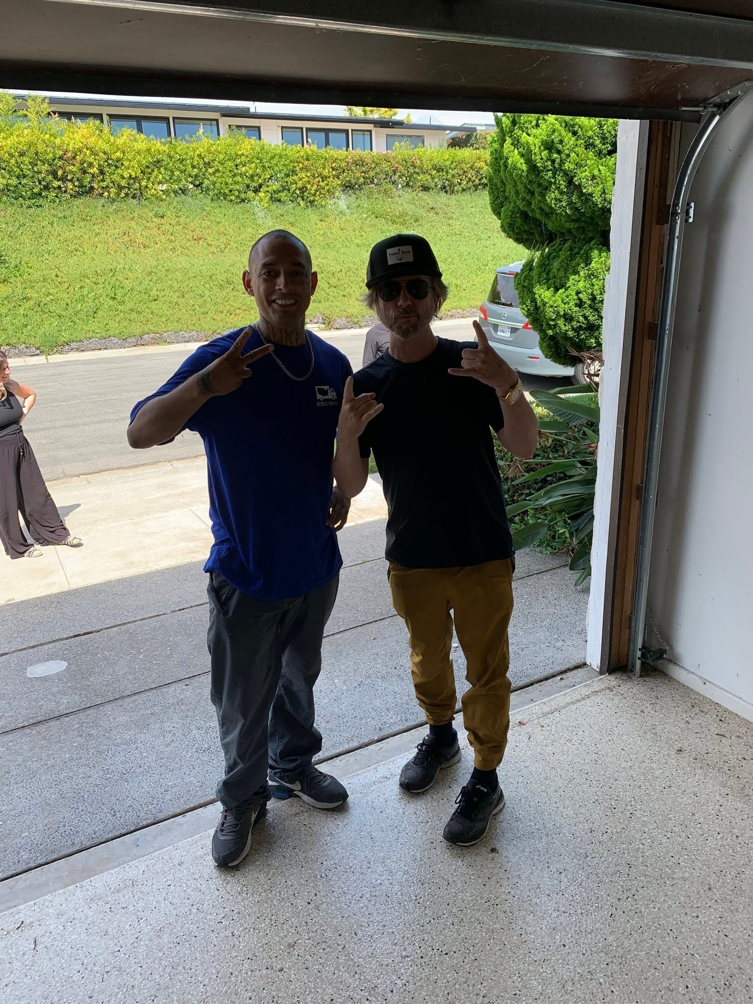 Two people standing in a garage doorway, smiling and making peace signs.