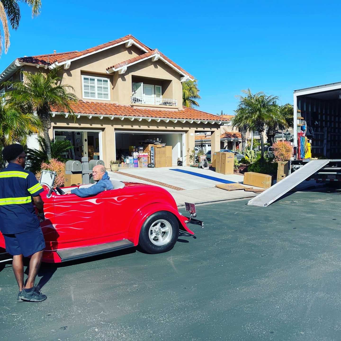 Red vintage convertible with driver outside a house, beside a moving truck ramp and a worker in a driveway