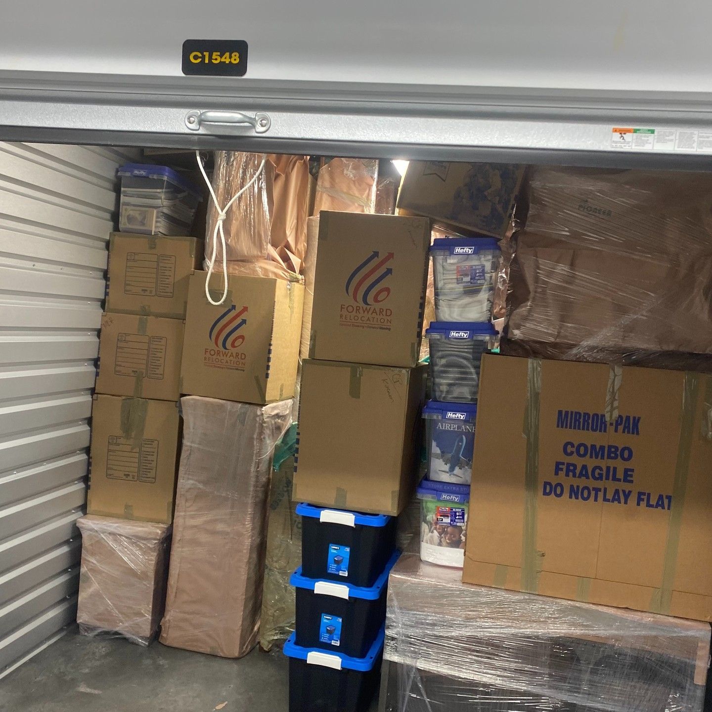 Packed storage unit with stacked cardboard boxes, blue bins, and wrapped packages on a metal floor.