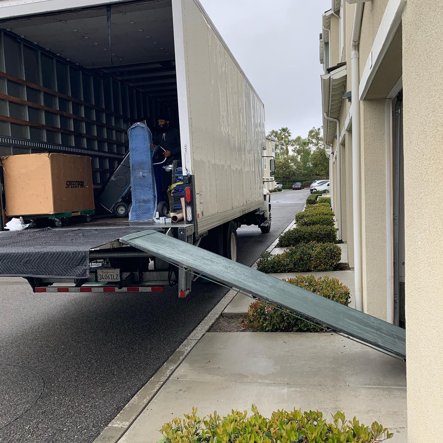 Moving truck with ramp extended onto a wet sidewalk beside a building.